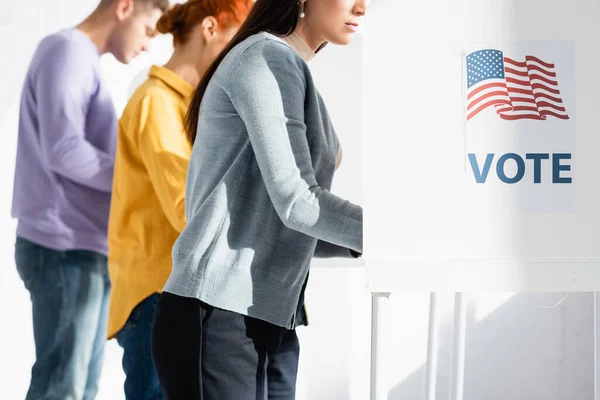Votantes en cabinas electorales con bandera americana e inscripción de voto sobre fondo borroso - foto de stock