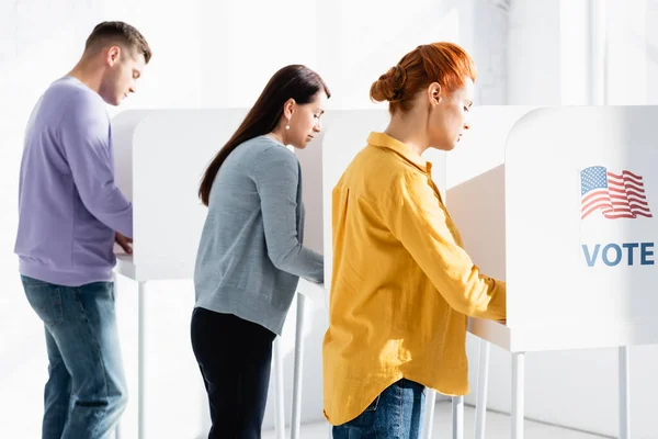 Electores en cabinas electorales con bandera americana y letras de voto sobre fondo borroso - foto de stock