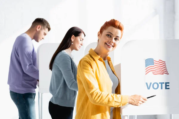 Sonriente mujer señalando con la pluma en la bandera americana y la inscripción de voto en la cabina de votación sobre fondo borroso - foto de stock