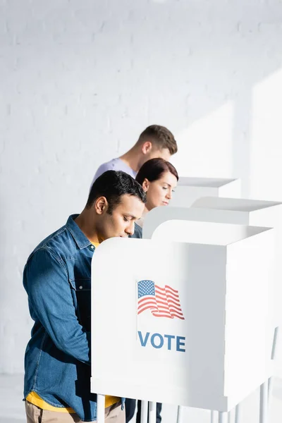 Multicultural voters in polling cabins with american flag and vote inscription on blurred background — Stock Photo