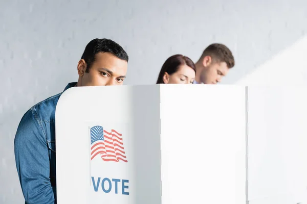 Indian man looking at camera from polling booth near multicultural electors on blurred background — Stock Photo