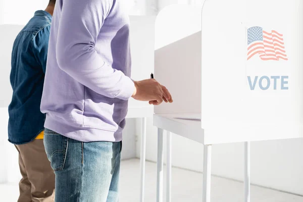 Vista ritagliata di uomini multiculturali in cabina elettorale con bandiera americana e iscrizione di voto — Foto stock