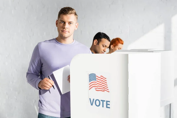 Man looking at camera while holding ballot near polling booth with american flag and vote lettering on blurred background — Stock Photo