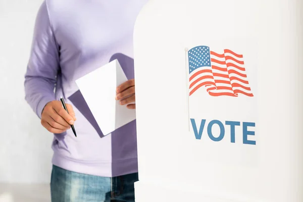 Cropped view of man holding ballot and pen near polling booth with american flag and vote inscription — Stock Photo