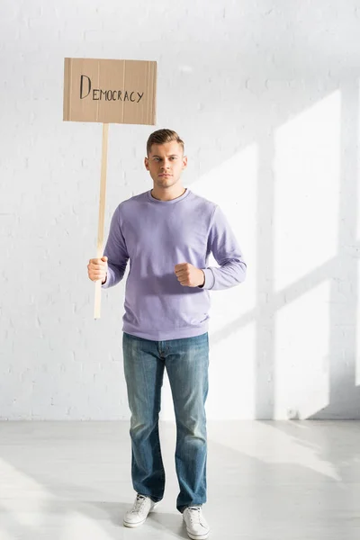 Ernsthafter Mann hält Plakat mit Demokratie-Inschrift an weiße Ziegelwand — Stockfoto