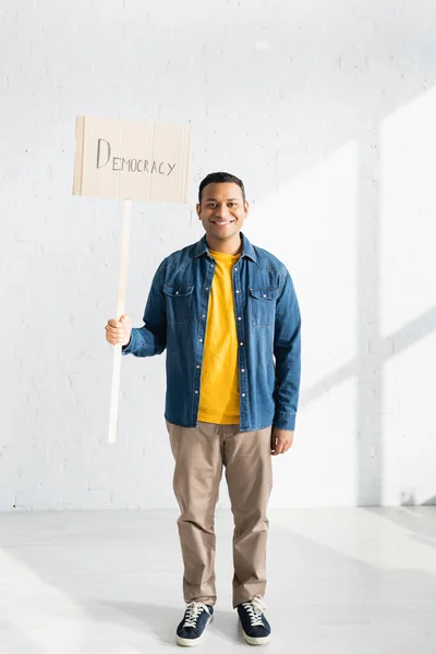 Smiling indian man holding placard with democracy lettering against white brick wall — Stock Photo