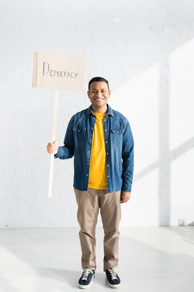 Smiling indian man holding placard with democracy lettering against white brick wall — Stock Photo