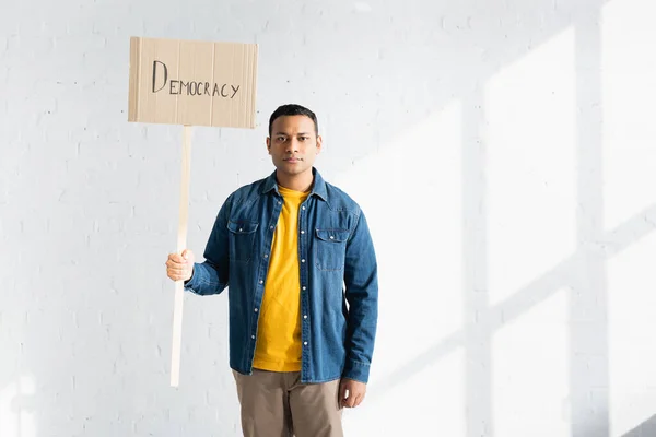 Serious indian man holding placard with democracy inscription against white brick wall — Stock Photo