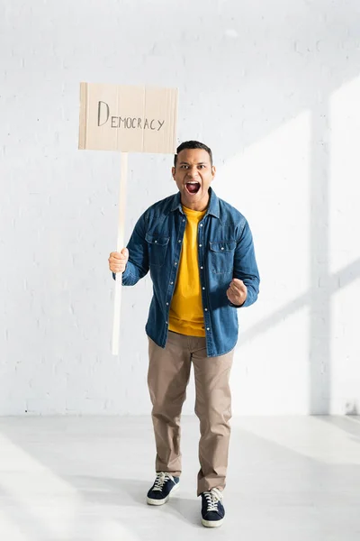 Aggressive indian man screaming while holding placard with democracy lettering against white brick wall — Stock Photo