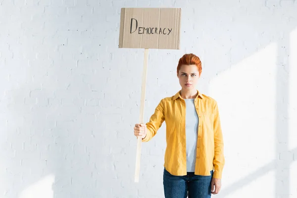 Mujer seria sosteniendo pancarta con las letras de la democracia contra la pared de ladrillo blanco - foto de stock