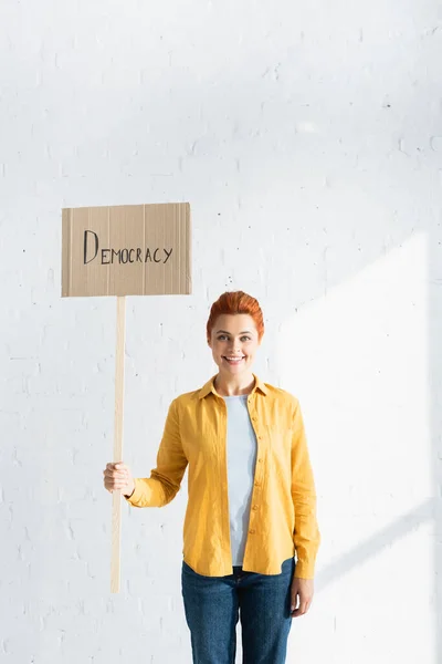 Femme souriante tenant une pancarte avec des lettres de démocratie contre un mur de briques blanches — Photo de stock