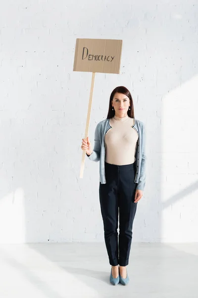 Serious woman holding placard with democracy inscription against white brick wall — Stock Photo