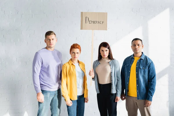 Serious multicultural people looking at camera while holding placard with democracy lettering — Stock Photo