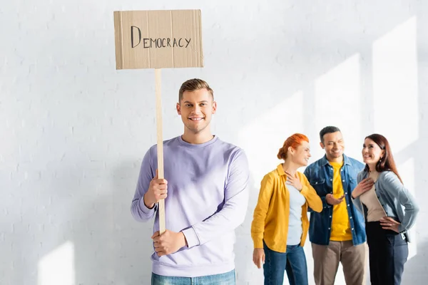 Felice uomo che tiene cartello con iscrizione democrazia mentre multiculturale persone che la pensano come parlare su sfondo sfocato — Foto stock