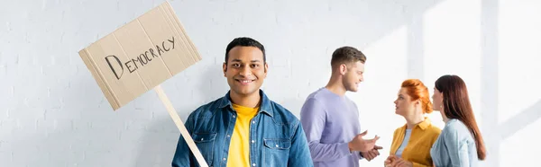 Hombre indio sonriente sosteniendo pancarta con letras de democracia cerca de personas multiculturales hablando en un fondo borroso, pancarta - foto de stock