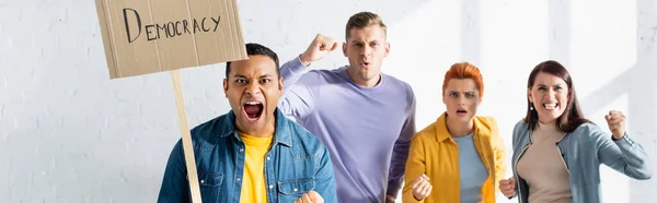 Aggressiver indischer Mann mit Plakat mit Demokratie-Schriftzug in der Nähe schreiender Gleichgesinnter auf verschwommenem Hintergrund, Transparent — Stockfoto