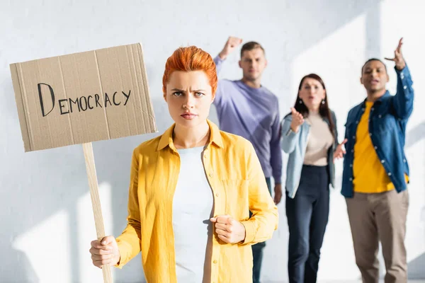 Mulher irritada segurando cartaz com inscrição democracia enquanto irritado pessoas multiculturais gesticulando em fundo turvo — Fotografia de Stock