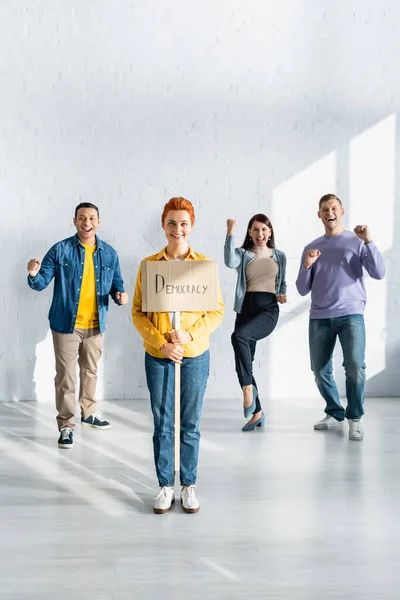 Mulher feliz segurando cartaz com democracia lettering perto de pessoas multiculturais felizes dançando no fundo — Fotografia de Stock