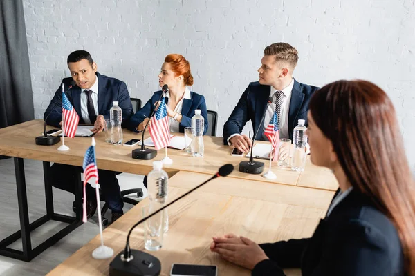 Políticos multiétnicos hablando durante el congreso del partido mientras están sentados en el escritorio con banderas de EE.UU. - foto de stock