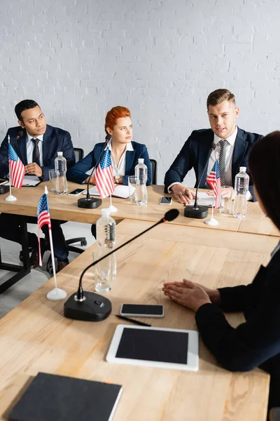 Multicultural politicians talking during party congress while sitting at desk with usa flags — Stock Photo