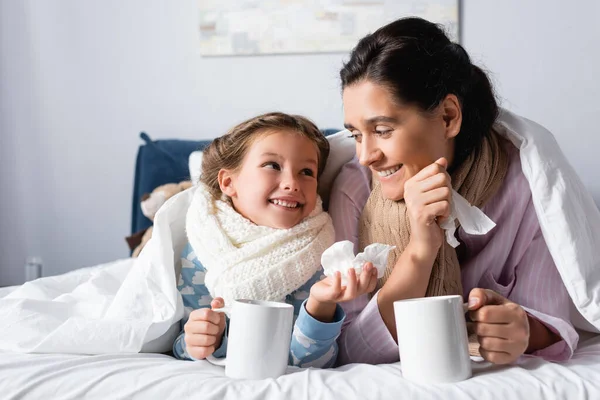 Madre e hija enfermas y alegres mirándose mientras sostienen tazas con bebida caliente en la cama - foto de stock