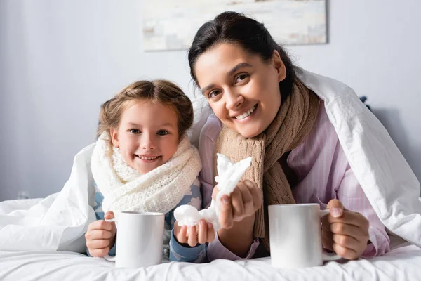 Mujer enferma y sonriente con un niño sosteniendo tazas de bebida caliente en la cama - foto de stock