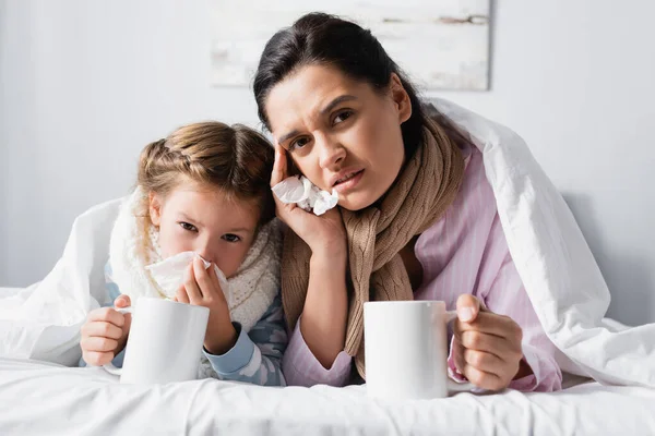 Upset mother with child looking at camera while holding warm drink in bed — Stock Photo
