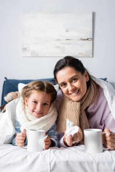 Cheerful mother and daughter looking at camera while lying in bed with cups of warm drink — Stock Photo