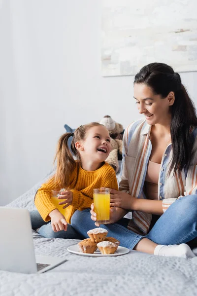 Feliz madre e hija mirándose mientras sostienen el jugo de naranja y ven películas en la computadora portátil - foto de stock