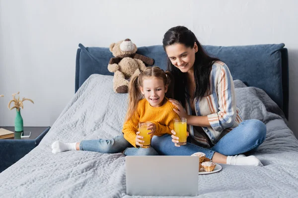 Cheerful woman with daughter holding orange juice while watching film on laptop — Stock Photo