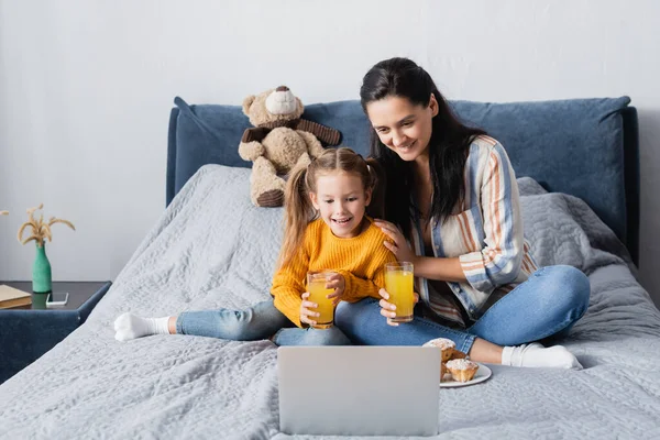 Donna allegra con figlia che tiene il succo d'arancia mentre guarda il film sul computer portatile — Foto stock