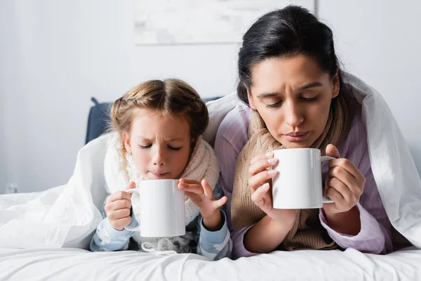 Madre e figlia malata che soffiano sul tè caldo mentre si trovano a letto — Foto stock