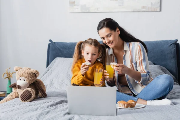 Возбужденный ребенок с счастливой матерью смотреть фильм на ноутбуке с апельсиновым соком и кексами — стоковое фото