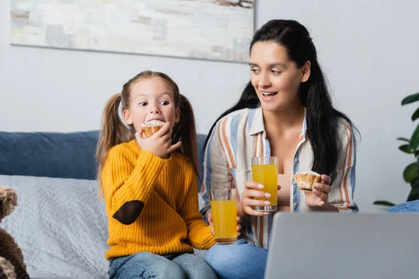 Daughter eating delicious muffin while watching film on computer with mother — Stock Photo