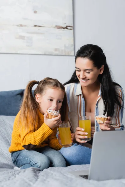 Happy mother looking at daughter eating muffin and watching movie on laptop — Stock Photo