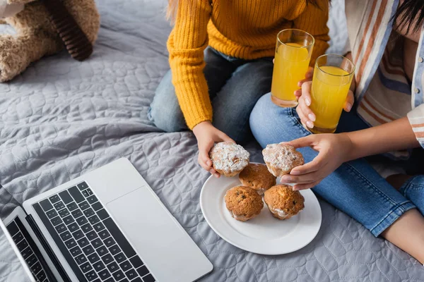 Cropped view of mom and child holding orange juice and muffins while watching movie on laptop — Stock Photo