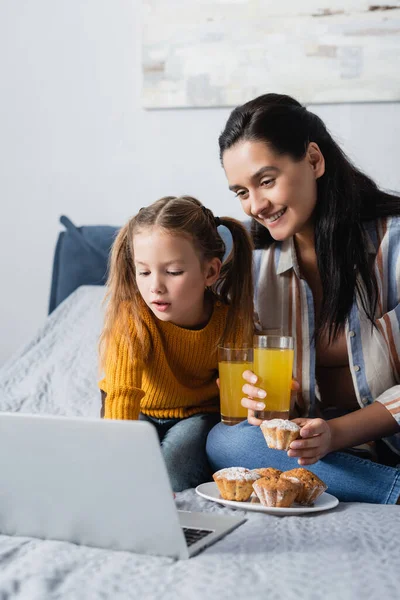 Madre e hija con magdalenas y jugo de naranja viendo película en el portátil - foto de stock