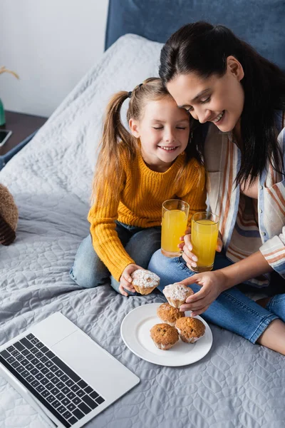Happy mother and daughter holding muffin and orange juice near laptop — Stock Photo