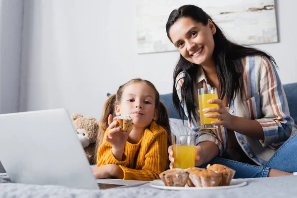 Happy woman and daughter looking at camera near muffins and orange juice while watching film on laptop — Stock Photo