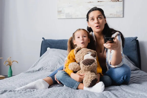 Excited mother and daughter watching tv while sitting on bed with teddy bear — Stock Photo