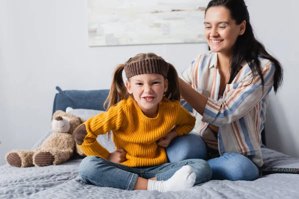 Grimacing girl in headband looking at camera while sitting near mother on bed — Stock Photo