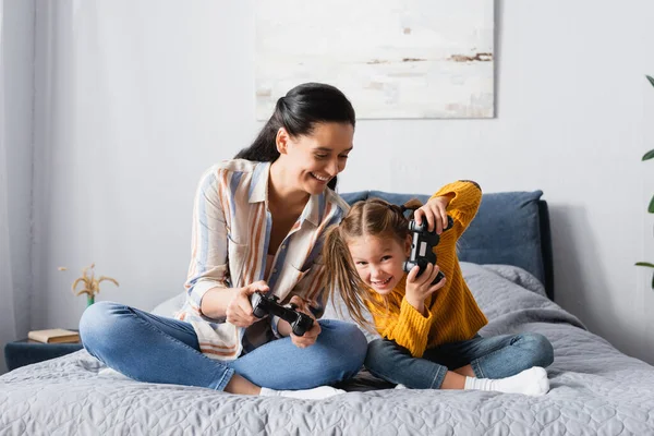 KIEW, UKRAINE - 15. SEPTEMBER 2020: Fröhliche Mutter und Kind spielen Videospiel, während sie mit gekreuzten Beinen auf dem Bett sitzen — Stockfoto