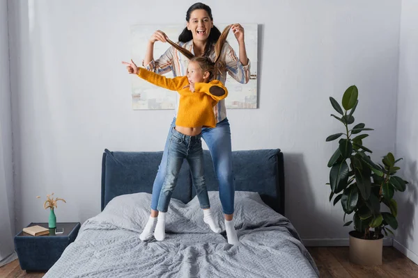 Excited mother holding hair of daughter while she pointing with finger and jumping on bed — Stock Photo