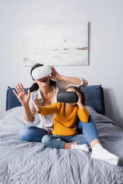 Mãe e filho usando fones de ouvido vr enquanto sentados na cama e gesticulando — Fotografia de Stock