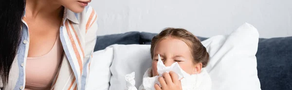 Mujer sentada cerca de hija enferma estornudando en servilleta de papel mientras está acostada en la cama, pancarta - foto de stock