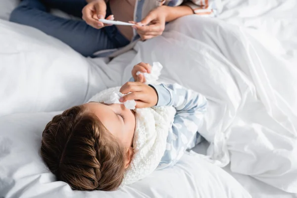 Vista aérea de la mujer mirando el termómetro cerca de la hija enferma acostada en la cama con servilleta de papel - foto de stock