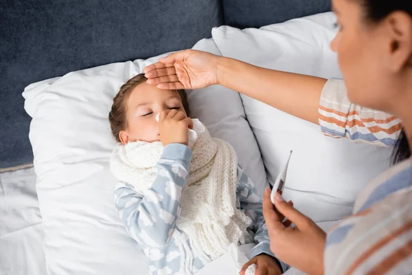 Niño enfermo limpiando la nariz con servilleta de papel mientras la madre sostiene el termómetro y toca su frente en un primer plano borroso - foto de stock
