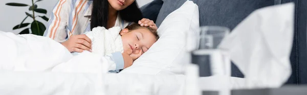 Madre toccando figlia malata dormire a letto vicino a un bicchiere d'acqua in primo piano sfocato, banner — Foto stock