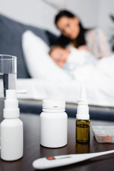 Garrafas com medicamentos na mesa de cabeceira perto de criança doente deitada na cama com a mãe no fundo turvo — Fotografia de Stock