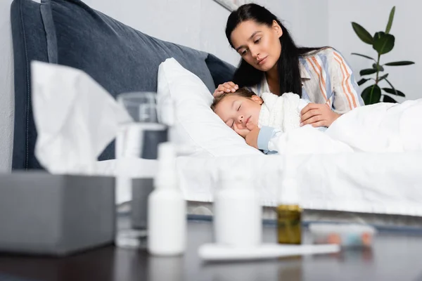 Мать обнимает больную дочь спит в постели рядом с прикроватным столиком с лекарствами на размытом переднем плане — стоковое фото